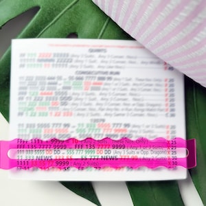 May include: A white card with printed text is held in place by a bright pink, scalloped-edge plastic holder. The card is partially obscured, but the text includes numbers and abbreviations. A green leaf and pink striped fabric are in the background.