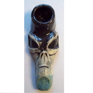 May include: A ceramic alien head pipe with black and white glaze. The pipe has a large bowl and a small mouthpiece.