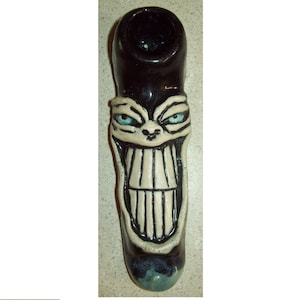 May include: A black and white ceramic pipe with a grinning face design. The pipe has a wide mouth and a small bowl. The face is painted with white details, including the eyes, teeth, and nose.