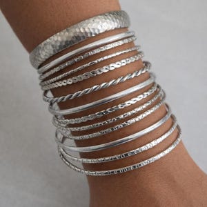 May include: A collection of silver bangles and bracelets adorns a wrist. The bracelets vary in design, including hammered, twisted, and patterned styles. The bangles are made of a shiny, metallic material.