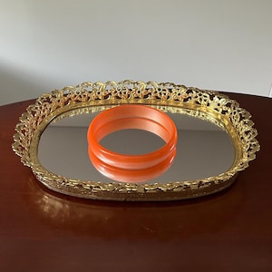 May include: A bright orange bangle bracelet rests on a mirrored oval tray. The tray features a gold-toned metal rim with an intricate, openwork floral pattern along the edge, reflecting the orange bracelet on its smooth, reflective surface.