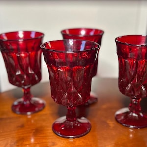 May include: A set of four deep red glass goblets featuring a faceted, diamond-like pattern on the exterior. Each goblet has a wide bowl, a short stem, and a circular base, all crafted from translucent ruby-colored glass with a reflective finish.