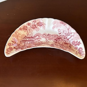 May include: A crescent-shaped ceramic dish featuring a maroon transferware pattern. The design depicts a landscape with a house, a bridge, a sailboat on water, and various trees and floral elements against a cream-colored background.