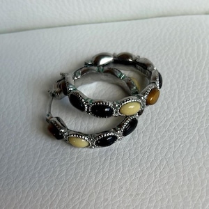 May include: A pair of silver hoop earrings. Each earring is adorned with a series of oval-shaped stones in black, cream, and brown. The earrings are displayed on a white surface.