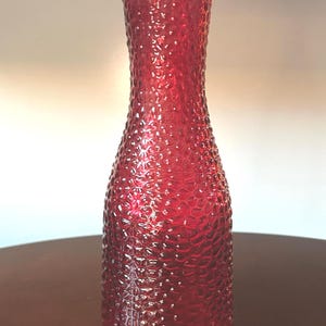 May include: A tall, cylindrical red glass vase featuring a textured, dimpled surface pattern throughout its exterior. The vessel has a narrow neck that flares slightly at the top rim, creating a distinct silhouette with a wide base and a smooth opening.