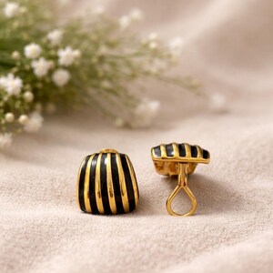 May include: A pair of gold-toned earrings with a black and gold striped design. The earrings have a rectangular shape with a rounded top and a post-back closure. The background is a soft, neutral color.