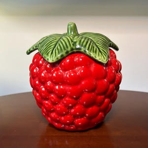 May include: A ceramic jar shaped like a bright red raspberry with a textured surface mimicking the fruit's drupelets. The lid is designed as a green stem with detailed leaves, featuring a glossy finish that covers the entire piece.