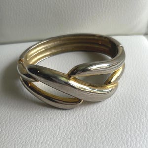 May include: A gold and silver-toned metal bracelet with an interlocking design. The bracelet has a hinged clasp and a polished finish. The design features a series of overlapping, curved elements, creating a stylish accessory.