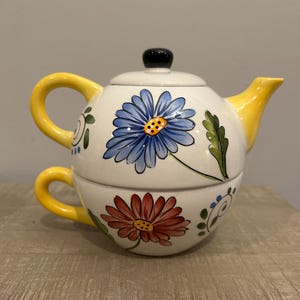 May include: A white ceramic teapot and cup set with a yellow handle and floral designs. The teapot has a blue flower with a yellow center and green leaves. The cup has a red flower with a yellow center.