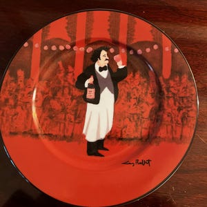 May include: A red ceramic plate with a black rim features a painting of a man in a white shirt and black vest holding a bottle of wine and a glass of wine. The man is surrounded by a crowd of people in a red and black setting. The plate is signed "Eric Pillot" in the bottom right corner.