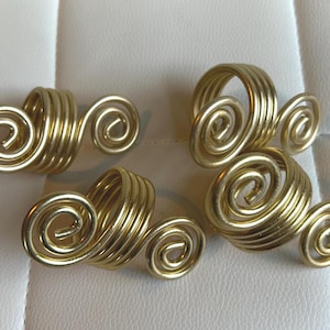May include: Four gold-colored napkin rings. Each ring features a cylindrical base with a spiral design on one side. The rings are made of a shiny, metallic material and are arranged on a white surface.