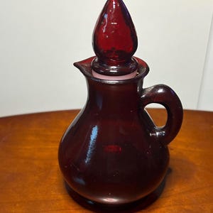 May include: A deep red glass cruet with a rounded body, a small handle on the side, and a pointed glass stopper. The vessel features a short spout for pouring and a smooth, translucent finish that allows light to pass through the dark red glass.