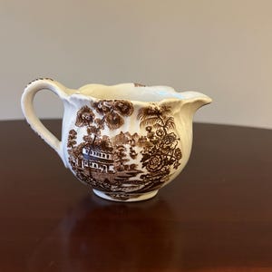 May include: A cream-colored ceramic creamer featuring a brown transferware pattern depicting a landscape with buildings, trees, and foliage. The creamer has a scalloped rim, a curved handle, and a small pouring spout on the opposite side.