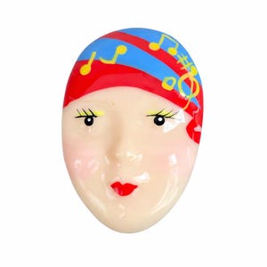 May include: A whimsical face-shaped brooch with a red and blue striped head covering adorned with yellow musical notes. The face has rosy cheeks, red lips, and delicate eyelashes. A unique accessory.