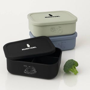 May include: Three-tiered lunch boxes in black, sage green, and blue. The top box is black with the text "MURKYOWL" and a white owl logo. The bottom box has a turkey graphic. A piece of broccoli sits nearby.