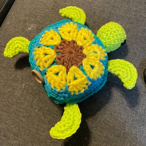 May include: A crocheted turtle toy with a bright yellow sunflower design on its shell. The turtle has green limbs and a brown center to the sunflower.