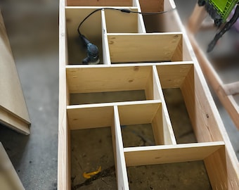Op de afbeelding: Een houten plank met meerdere vakken, gemaakt van onbehandeld hout. De plank is leeg, behalve voor een stroomkabel en een klein elektrisch gereedschap.