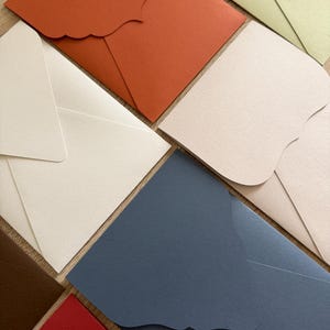 Könnte beinhalten: Eine Auswahl an farbenfrohen Umschlägen in verschiedenen Farbtönen, darunter Rot, Orange, Blau und Creme. Die Umschläge haben ein wellenförmiges Klappendesign und sind auf einer Holzoberfläche angeordnet. Das Bild zeigt Schreibwaren.