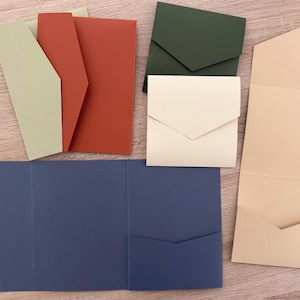 May include: A collection of coloured paper envelopes and cardstock in various shapes. Colours include sage green, rust, dark green, cream, navy blue, and tan. The envelopes have a triangular flap closure, and the cardstock is folded.