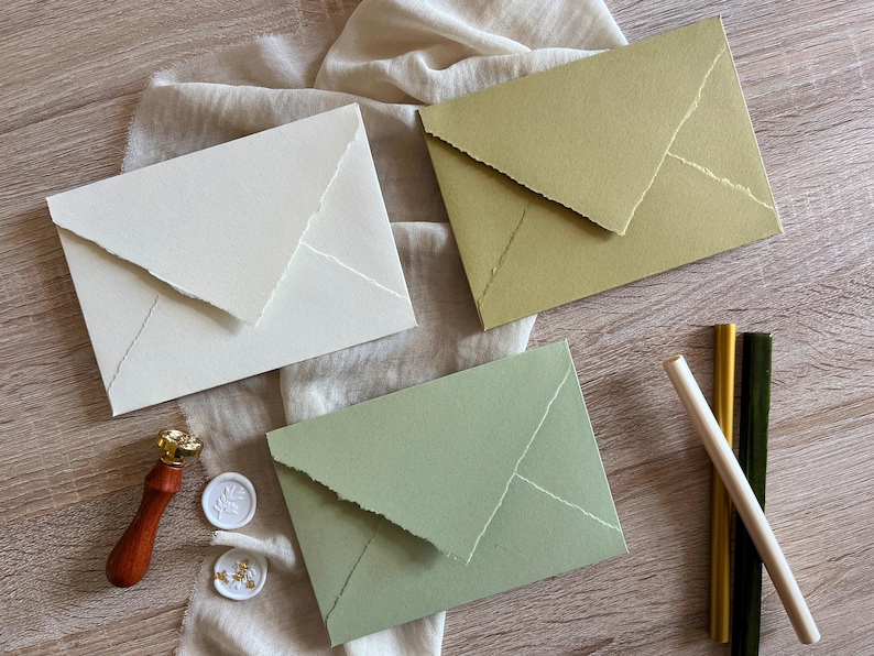 May include: Three envelopes with a textured paper finish. The envelopes are in white, green, and a light brown color. The envelopes have a flap on the top and are folded in half. The envelopes are on a wooden surface.