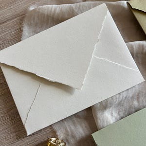 May include: A white envelope with a textured surface and a jagged edge design. The envelope is lying on a beige fabric surface.