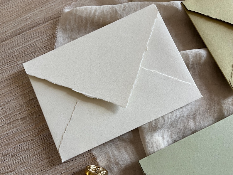 May include: A white envelope with a textured surface and a jagged edge design. The envelope is lying on a beige fabric surface.