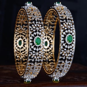 May include: A pair of gold bangles featuring an intricate openwork filigree design encrusted with numerous small clear stones. Each bangle is accented with a central oval-shaped green gemstone surrounded by a halo of small clear stones.