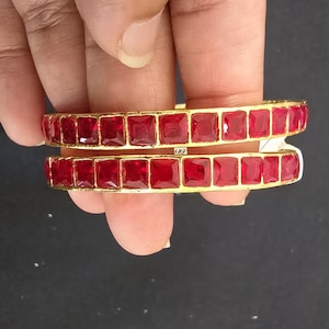 May include: A pair of gold-toned bangles featuring a continuous row of square-cut, deep red faceted stones set in a metallic frame. The bracelets have a structured, uniform design with a polished finish, showcasing a vibrant crimson color palette.
