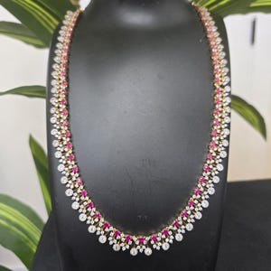May include: A gold-toned necklace and matching earrings set. The necklace features alternating white and ruby-colored stones. The earrings have a similar design. The jewelry is displayed on a black mannequin.