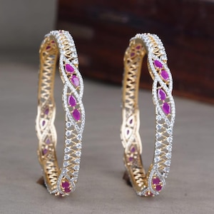 May include: A pair of gold-toned bangles featuring a twisted, openwork design. Each bangle is encrusted with clear, sparkling stones and accented with teardrop-shaped, vibrant pink stones arranged in a repeating pattern along the center.