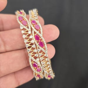 May include: Two gold bangles featuring a lattice design encrusted with clear white stones and accented with marquise-cut pink stones. The metal structure is intricate, with the stones set in a repeating pattern along the entire circumference.