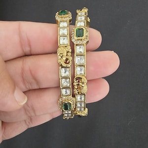 May include: Two gold-toned bangles featuring square-cut clear stones and rectangular green stones. Each bangle is accented with a small gold elephant motif and a border of tiny clear stones, creating a detailed and structured metal design.
