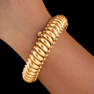May include: A gold-colored bracelet with a ribbed design and small, round accents. The bracelet is approximately 7 inches in length and is worn on a wrist against a black background. The jewelry piece has a classic, elegant appearance.