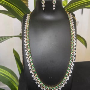 Puede incluir: Un conjunto de collar y pendientes dorados exhibido en un maniquí negro. El collar presenta piedras preciosas blancas y verdes alternadas. Los pendientes tienen un diseño similar. La joyería se presenta sobre un fondo de follaje verde.