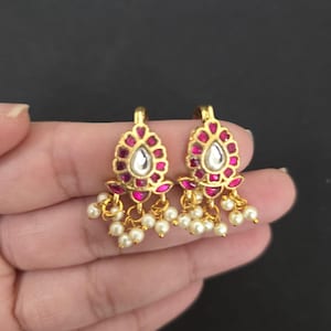 May include: A pair of gold-toned earrings with a teardrop-shaped, clear stone center, surrounded by pink gemstones. Small white pearls dangle from the bottom of each earring, creating a delicate and elegant design.