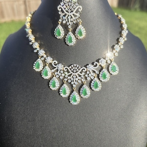 May include: A gold-toned necklace and matching headpiece set with clear crystals and teardrop-shaped green gemstones. The necklace and headpiece are displayed on a black mannequin head against a blurred outdoor background.