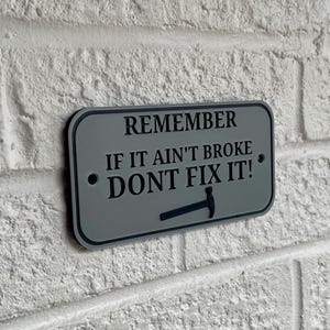 May include: A rectangular sign with rounded corners, featuring the text "REMEMBER IF IT AIN'T BROKE DONT FIX IT!" in black font. A small hammer graphic is below the text. The sign is gray with a black border and is mounted on a white brick wall.