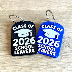 May include: Two graduation-themed keychains. One is black, the other blue. Both have a white graduation cap graphic and the text "CLASS OF 2026 SCHOOL LEAVERS" in white. Each keychain has a silver keyring attached.