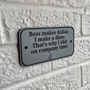 May include: A rectangular, grey sign with black text that reads "Boss makes dollar, I make a dime. That's why I shit on company time." The sign is mounted on a white brick wall.