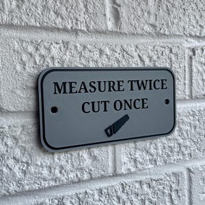 May include: A rectangular gray sign with rounded corners is mounted on a white brick wall. The sign reads "MEASURE TWICE CUT ONCE" in black text, with a saw graphic below. Two small holes are visible on the sign.