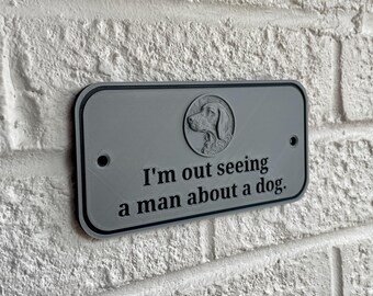 May include: A gray rectangular sign with rounded corners is mounted on a white brick wall. The sign features a dog silhouette and the phrase "I'm out seeing a man about a dog." The sign has a black border.