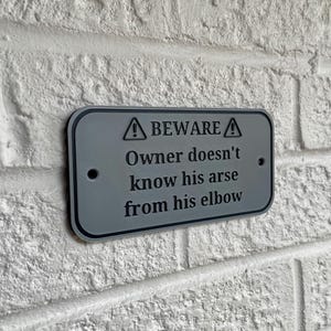 Puede incluir: Un letrero rectangular gris con borde negro est&aacute; montado en una pared de ladrillos blancos. El letrero dice "BEWARE" encima del texto "Owner doesn't know his arse from his elbow."