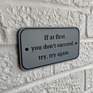 Puede incluir: Un letrero rectangular plateado con esquinas redondeadas est&aacute; montado en una pared de ladrillos blancos. El letrero presenta la frase "If at first you don't succeed, try, try again" en texto negro. El letrero tiene dos peque&ntilde;os agujeros.