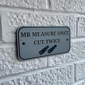 May include: A rectangular, gray sign with black trim and the text "MR MEASURE ONCE CUT TWICE" in black. Two saw icons are below the text. The sign is mounted on a white brick wall.