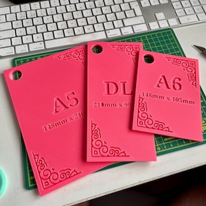 Peut inclure: Trois gabarits rectangulaires rose vif avec des motifs décoratifs dans les coins et des trous circulaires en haut. Les gabarits sont marqués A5, DL et A6 avec leurs dimensions respectives en millimètres imprimées sur la surface de chaque pièce.