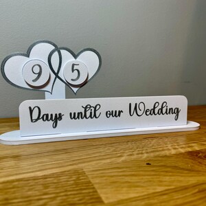 May include: White countdown sign with two interlocking hearts, each with a silver circle containing a number. The sign reads "Days until our Wedding".