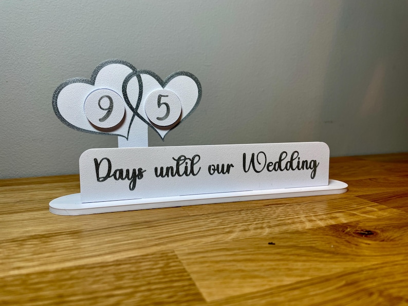 May include: White countdown sign with two interlocking hearts, each with a silver circle containing a number. The sign reads "Days until our Wedding".