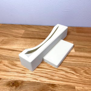 May include: White plastic stand with a curved slot for holding a video game cartridge. The stand is on a wooden surface.