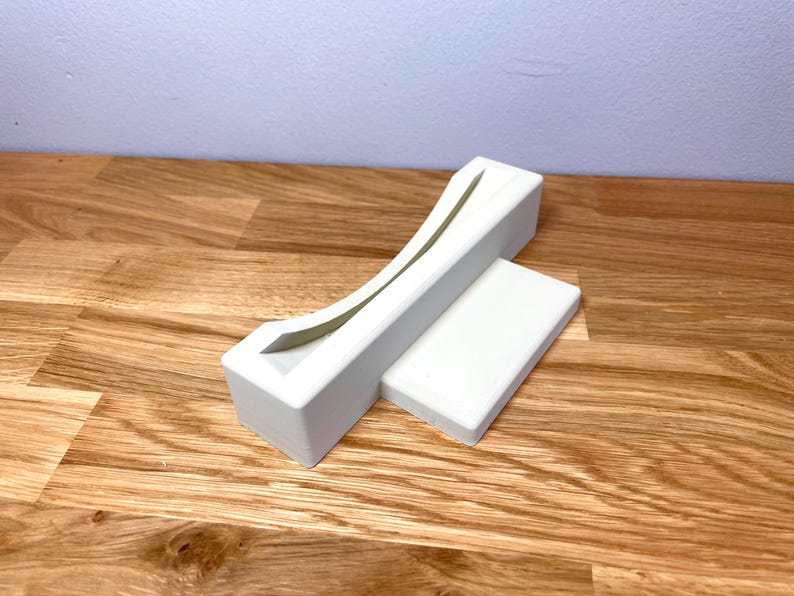 May include: White plastic stand with a curved slot for holding a video game cartridge. The stand is on a wooden surface.