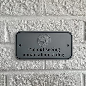 May include: A rectangular, gray metal sign with rounded corners is mounted on a white brick wall. The sign features a dog silhouette in a circle and the phrase, "I'm out seeing a man about a dog."